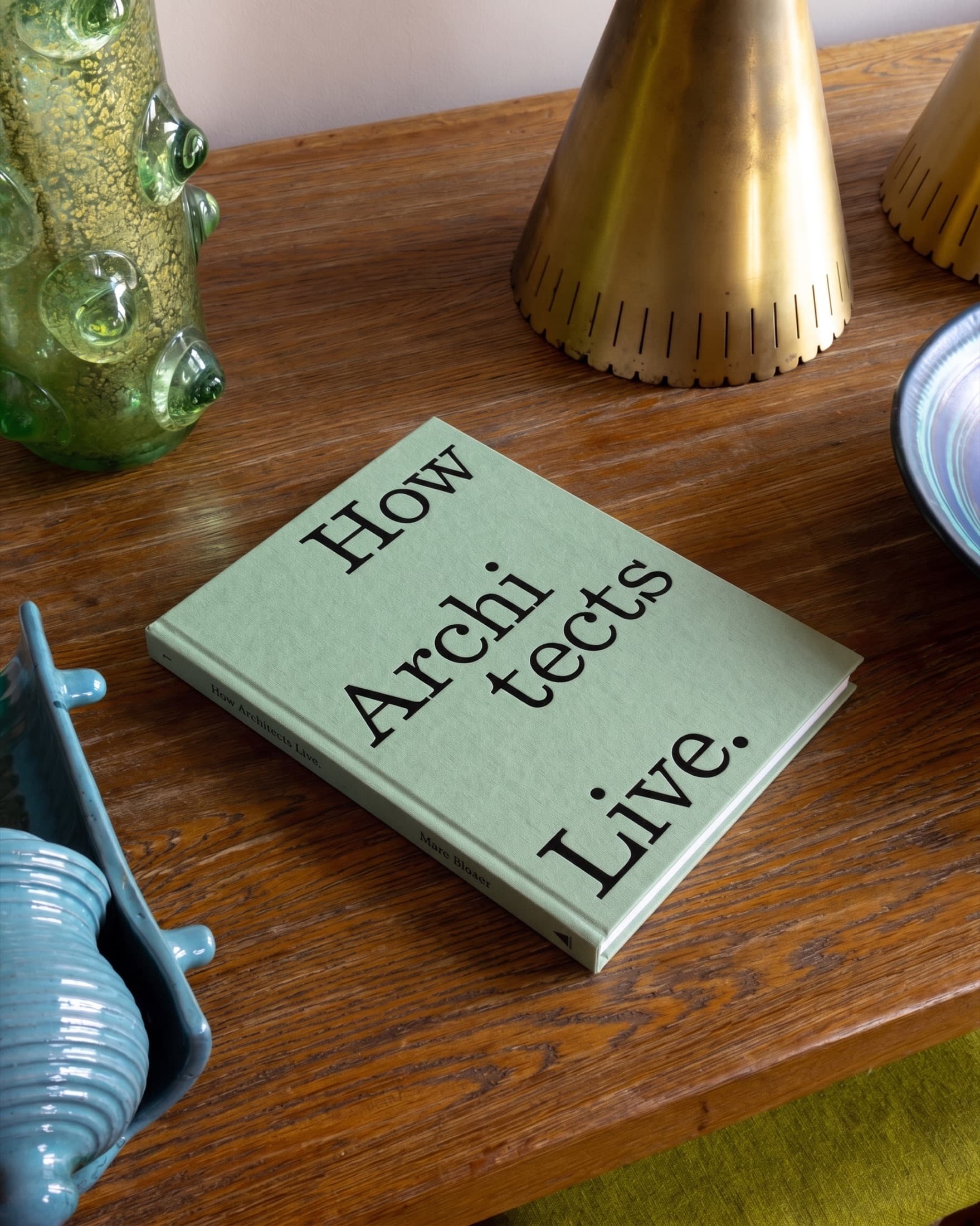 The book on a wooden sideboard among art glass and a brass lamp.