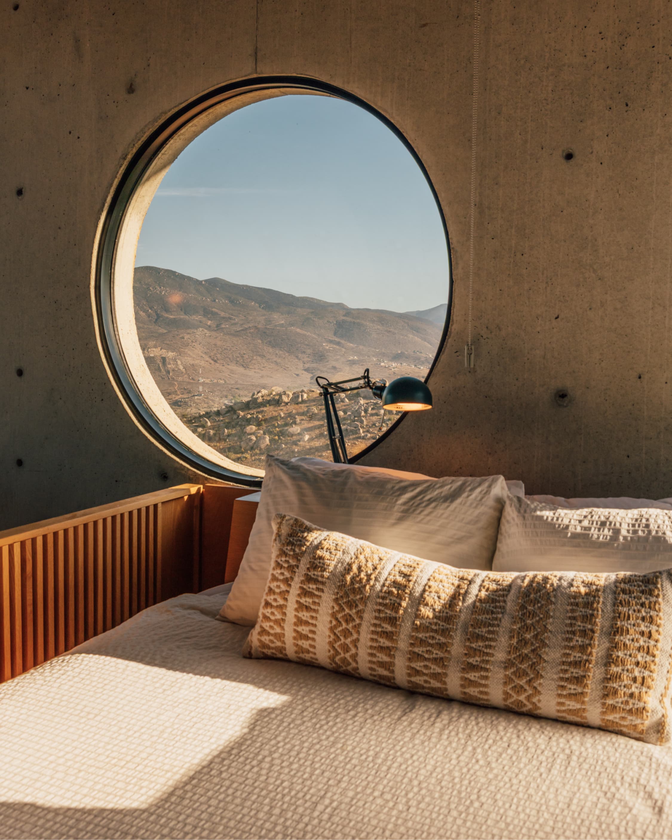 A bed beneath a large circular window framing an arid mountain valley in warm evening light.