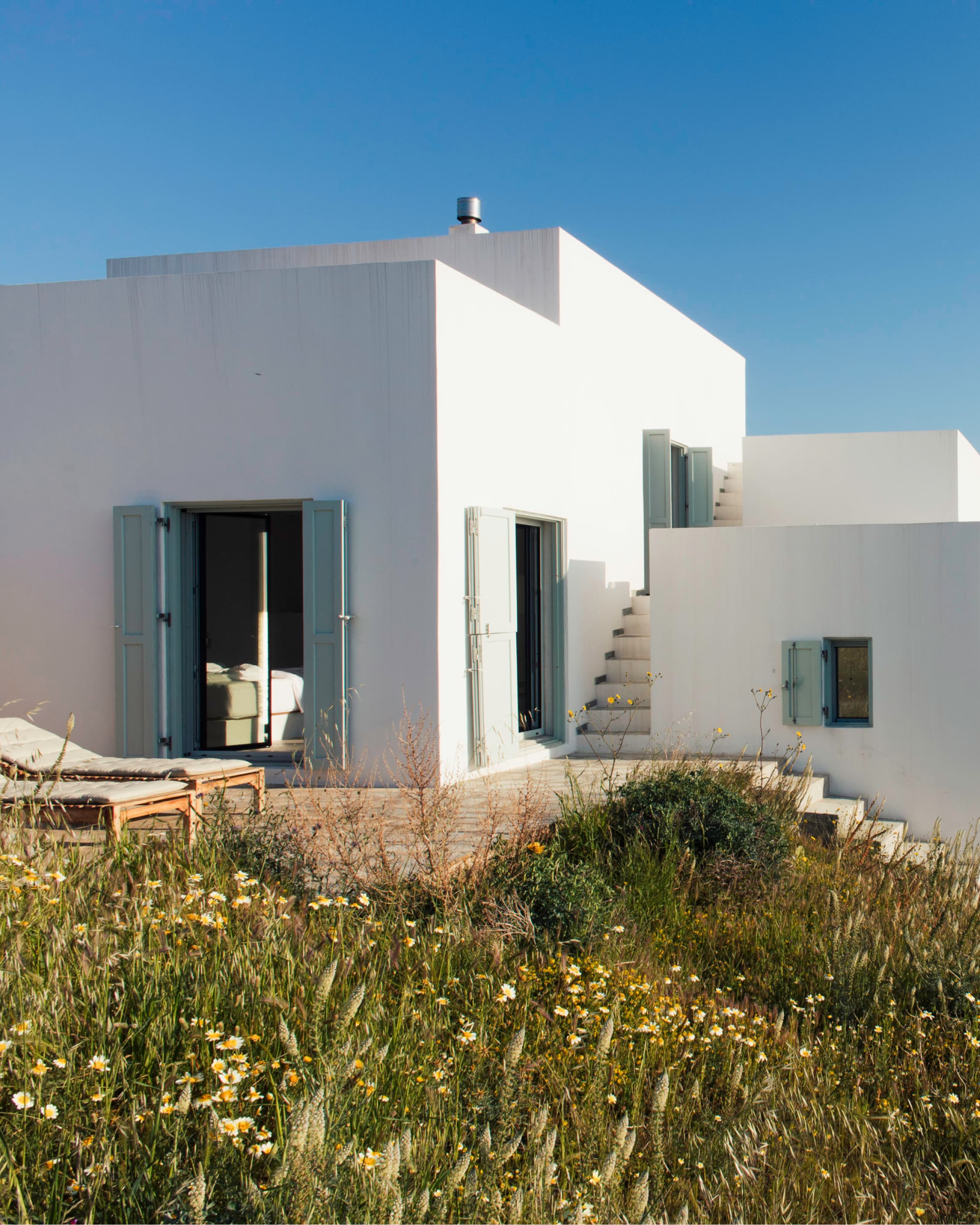 The whitewashed Cycladic house with sage shutters, external stairs, wildflowers on the slope.