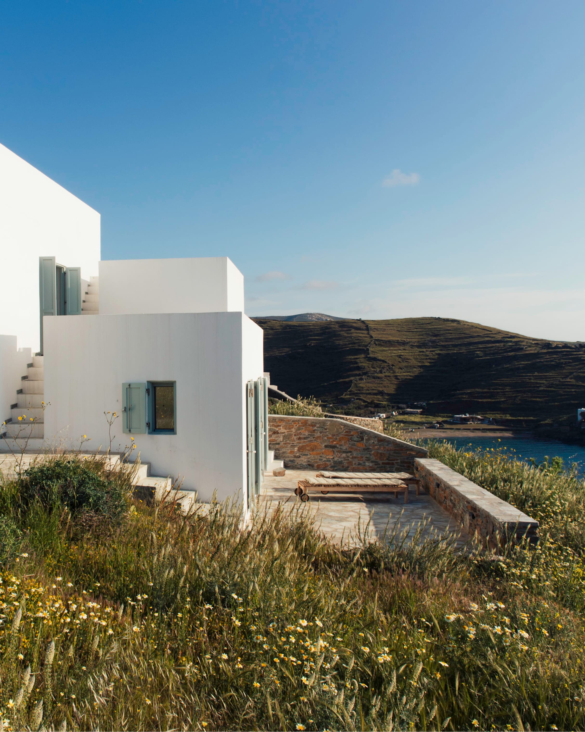 Stone terrace of the white island house, external stairs, a daybed, the bay and bare hills beyond.