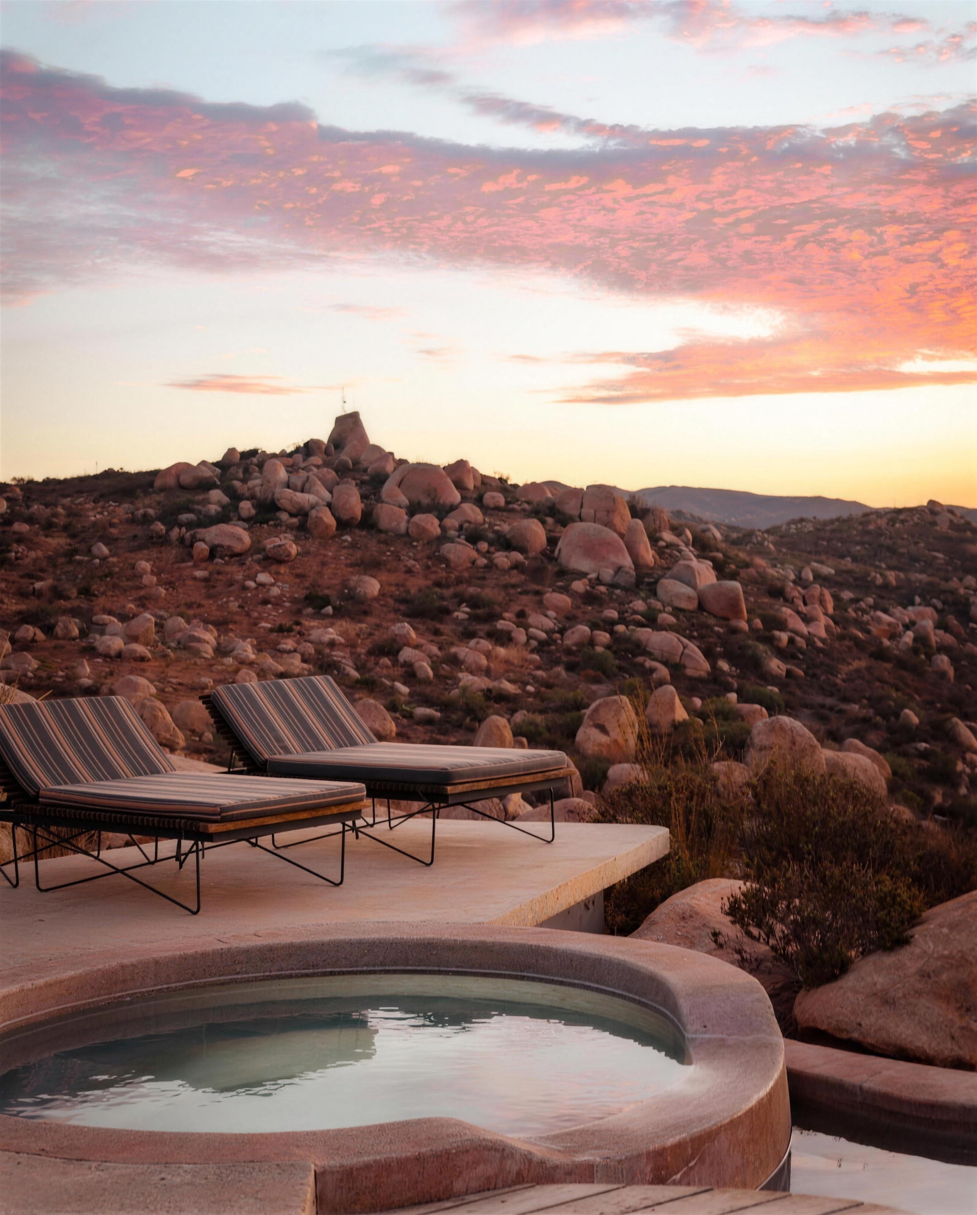 Two striped chaise lounges on a concrete terrace beside a round plunge pool, boulders and an arid hillside beyond, under a pink sunset sky.