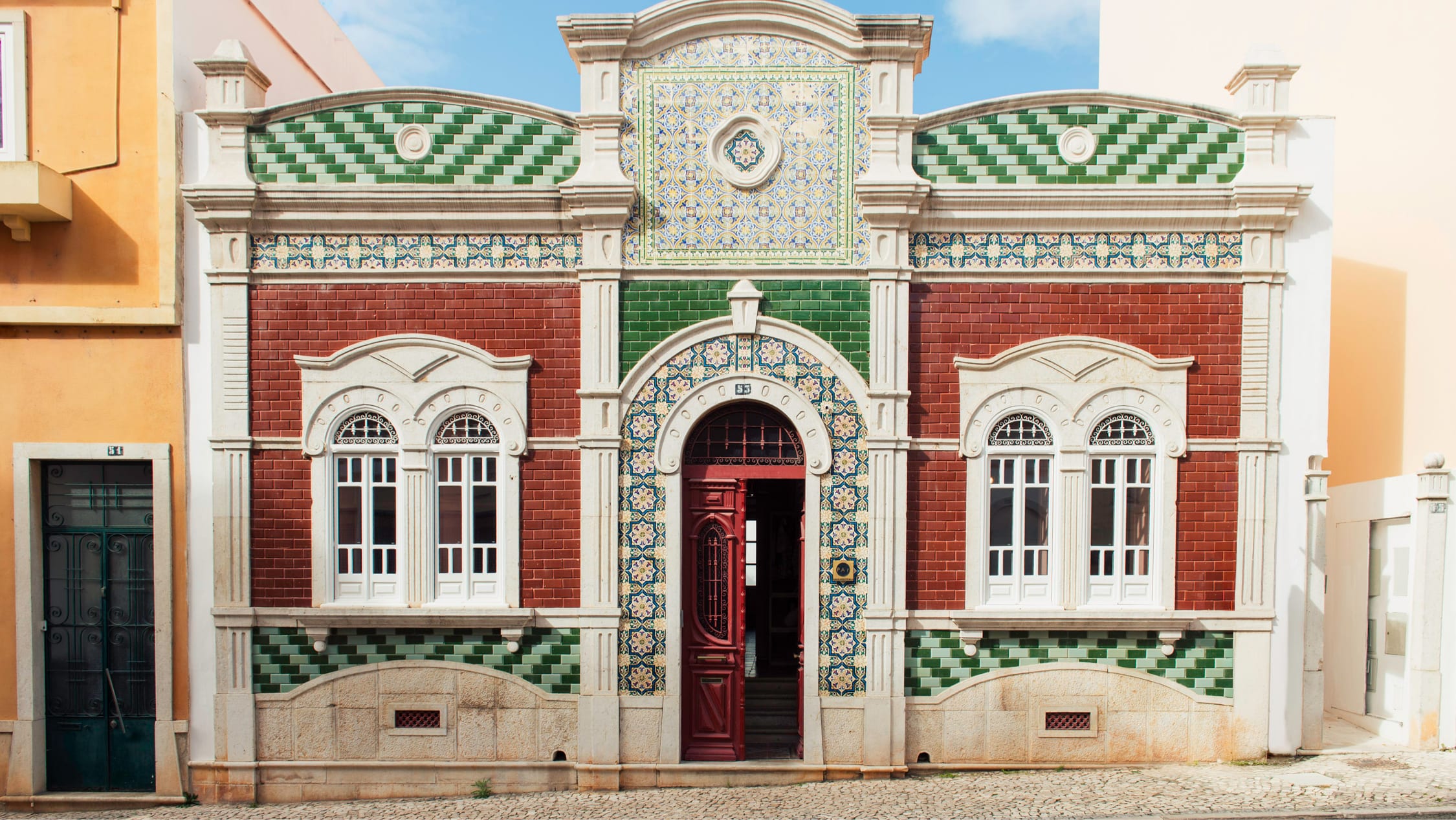 An ornate tiled townhouse façade — green and terracotta azulejos, white stucco, a deep red door standing open.