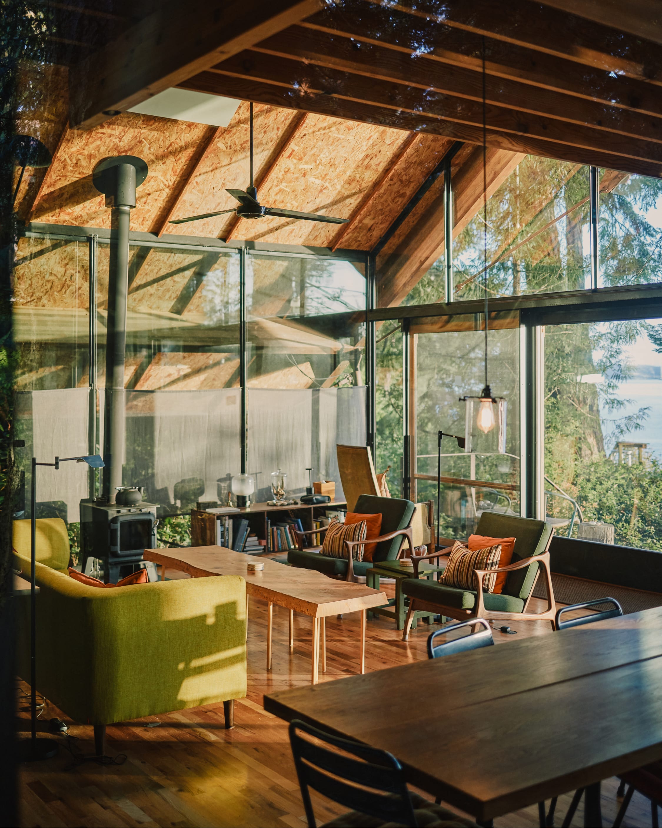 A glass-walled cabin living room with a timber A-frame ceiling, wood stove, mid-century chairs, forest and water beyond.