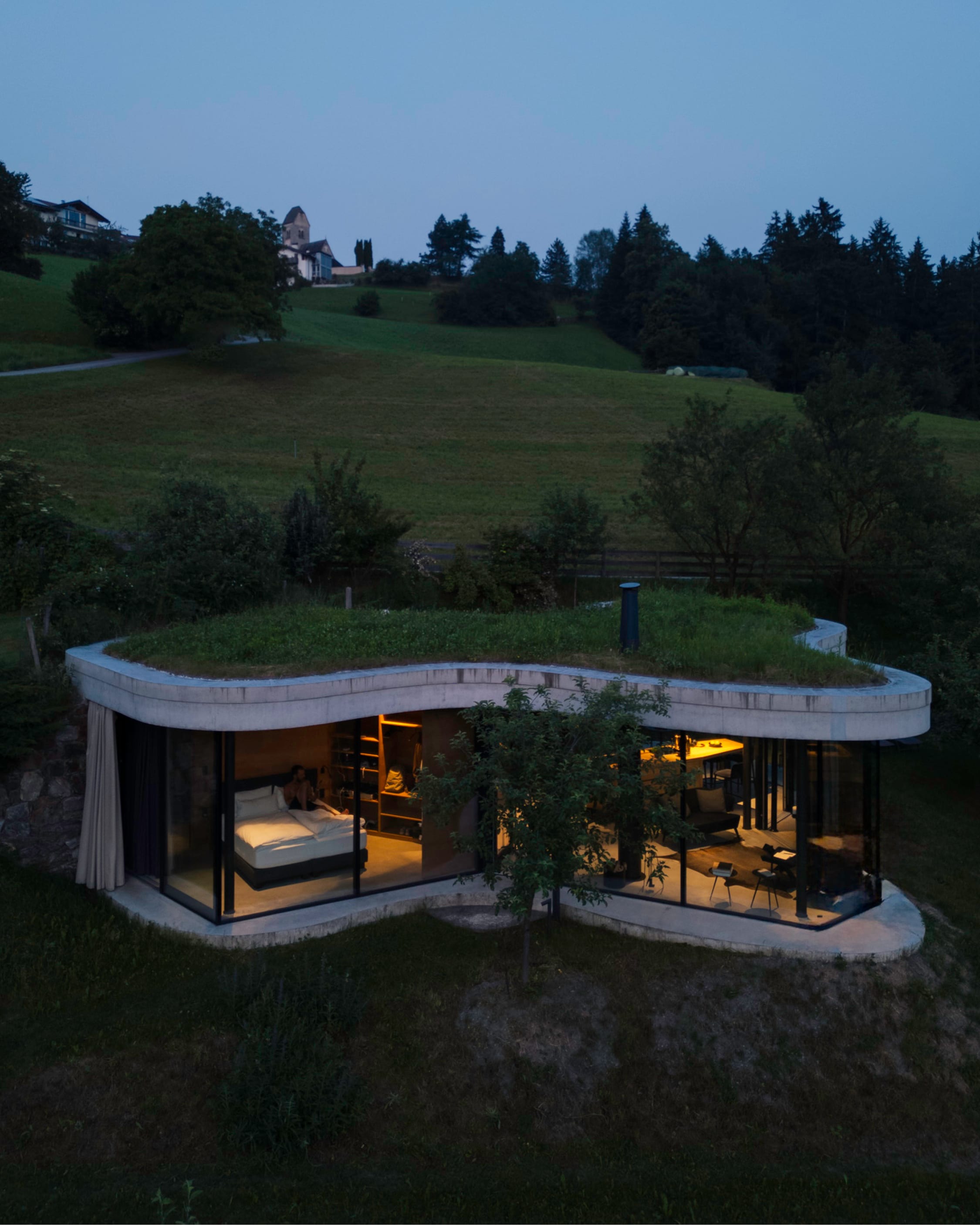 A curved, grass-roofed house set into an alpine hillside at dusk, glass walls glowing warm.