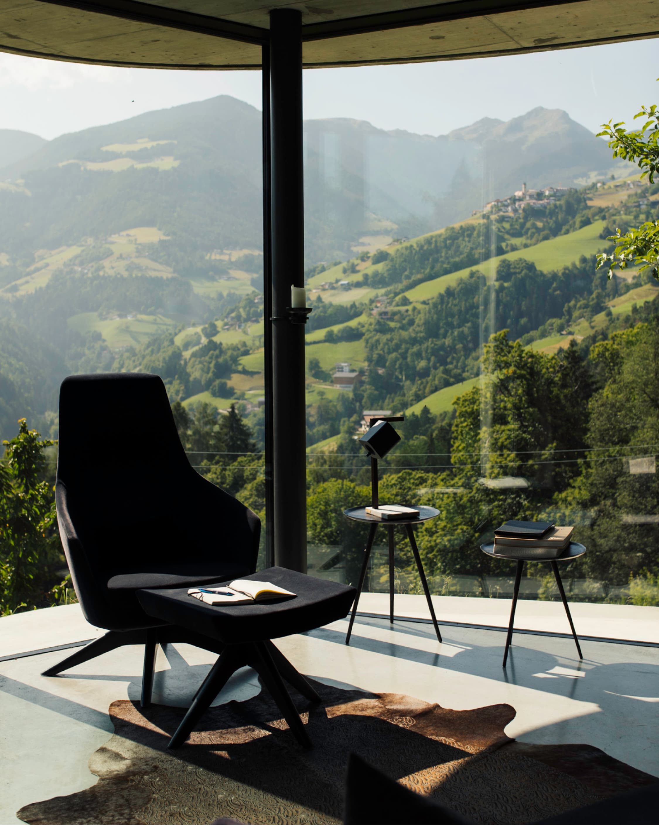 A black lounge chair on polished concrete, an open book on the seat, curved glass onto a green valley and hilltop village.