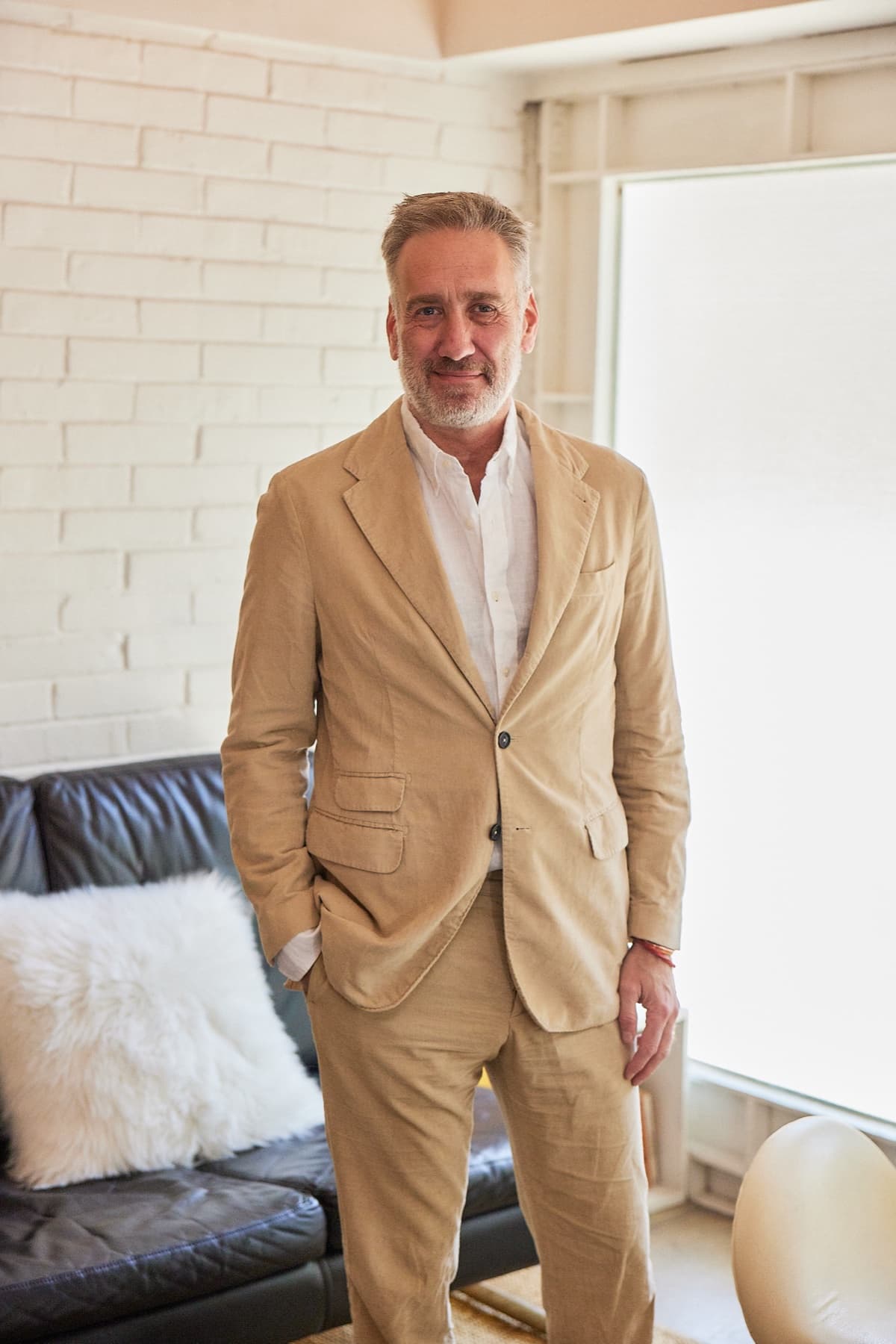 Marc Blazer in a camel corduroy suit, standing in a light-filled room with a black leather sofa.